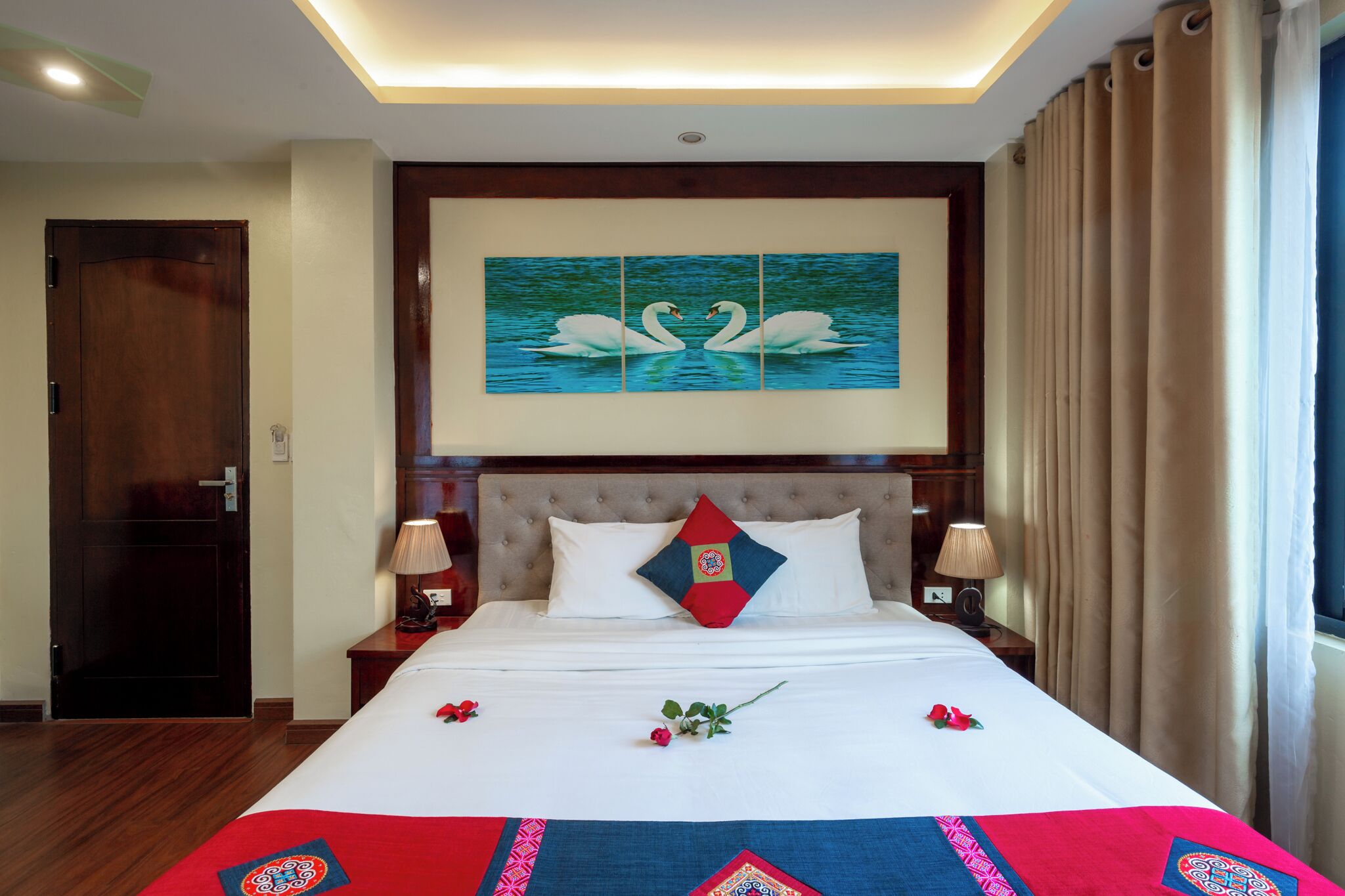 Una habitación de hotel con una cama grande, dos lámparas de noche, un cuadro colgado en la pared con cisnes, una colcha de edredón con estampado rojo y azul, y una puerta de madera.