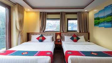 A hotel room with two beds, beige curtains, a nightstand with a lamp, and a painting on the wall.