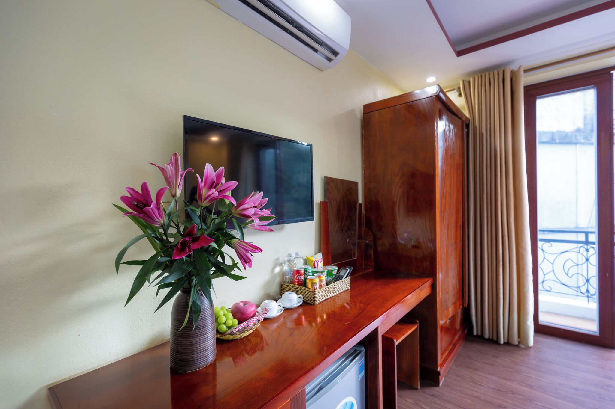 A hotel room with a wooden desk, a flat-screen TV, a vase of flowers, and a window with curtains.