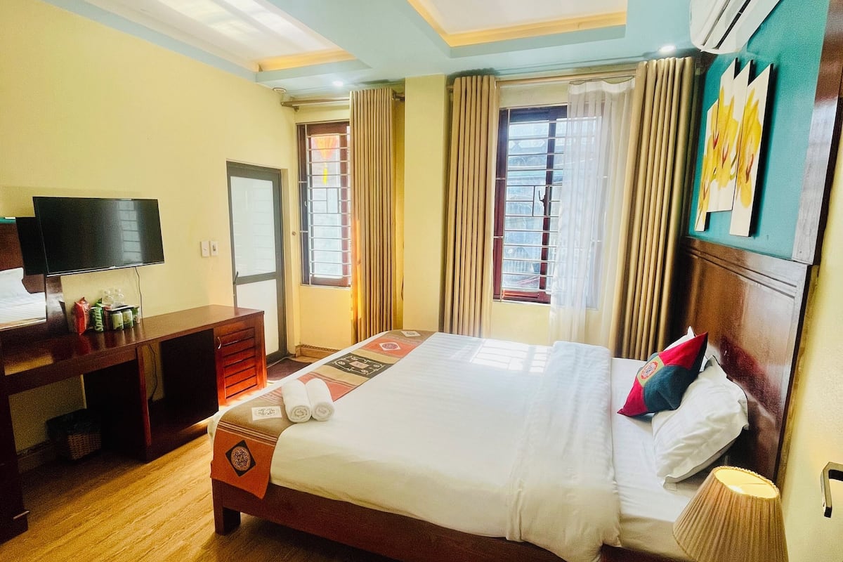 A hotel room with a large bed, wooden floor, a desk with a flat-screen TV, and a window with curtains.