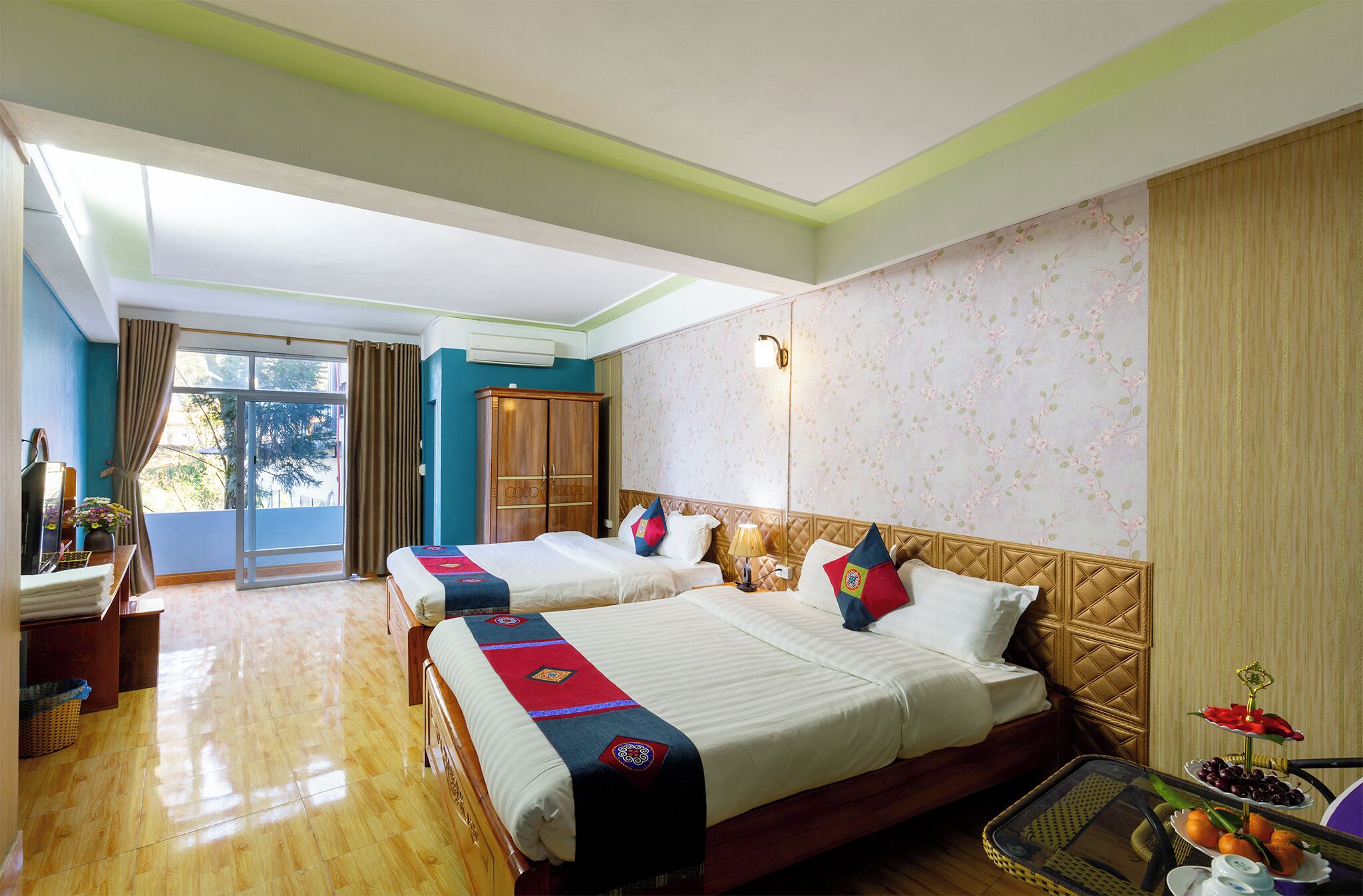 Une chambre d’hôtel avec deux lits, un bureau, une chaise, une petite table avec des fruits et une fenêtre avec des rideaux.