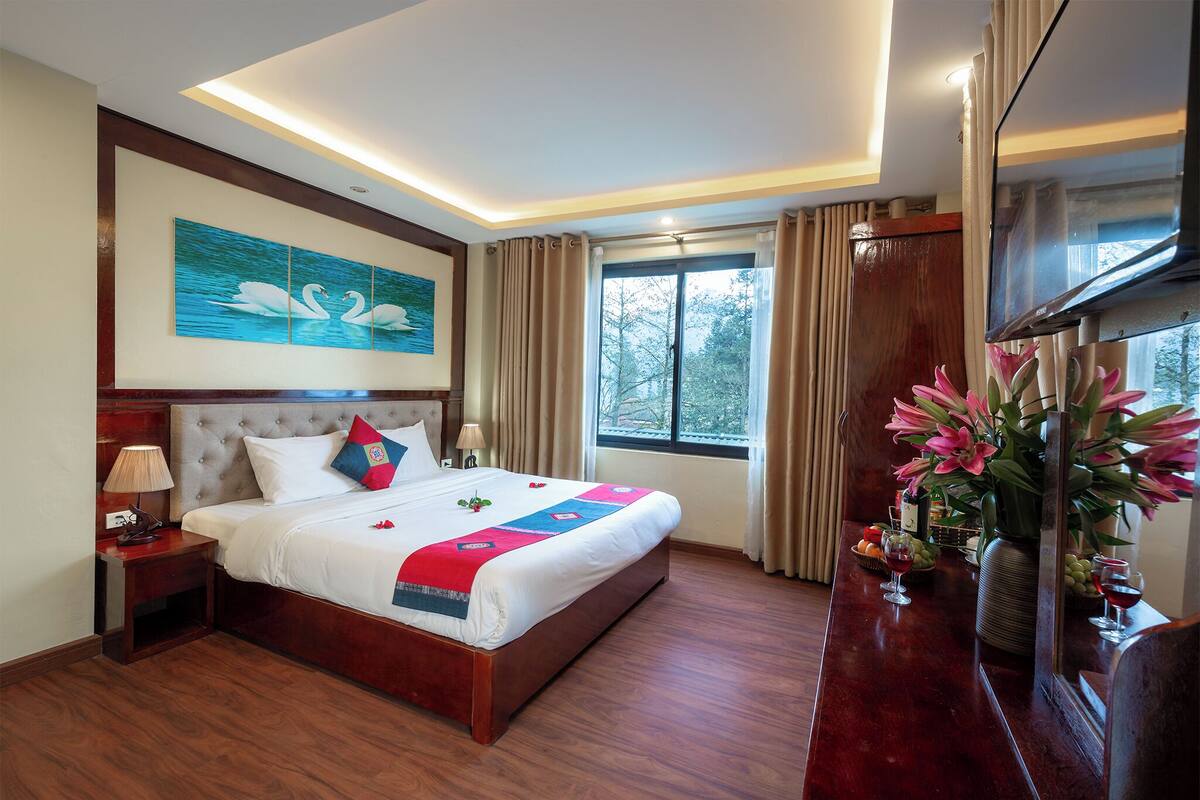 A hotel room with a large bed, a flat-screen TV, a nightstand, a bedside table, a vase with flowers, and a window with curtains.