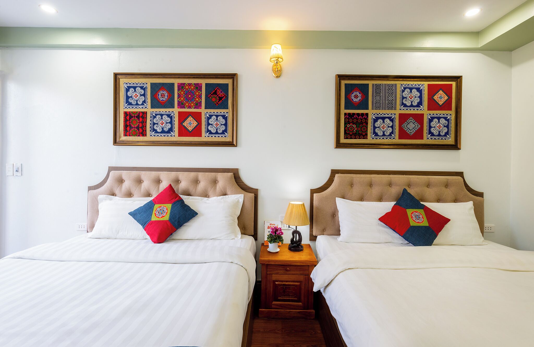 Habitación de hotel con dos camas, cada una con cabecero capitonado y ropa de cama blanca. Hay dos adornos enmarcados en la pared sobre las camas, una mesita de noche con una lámpara y una planta pequeña entre las camas, y piso de madera.