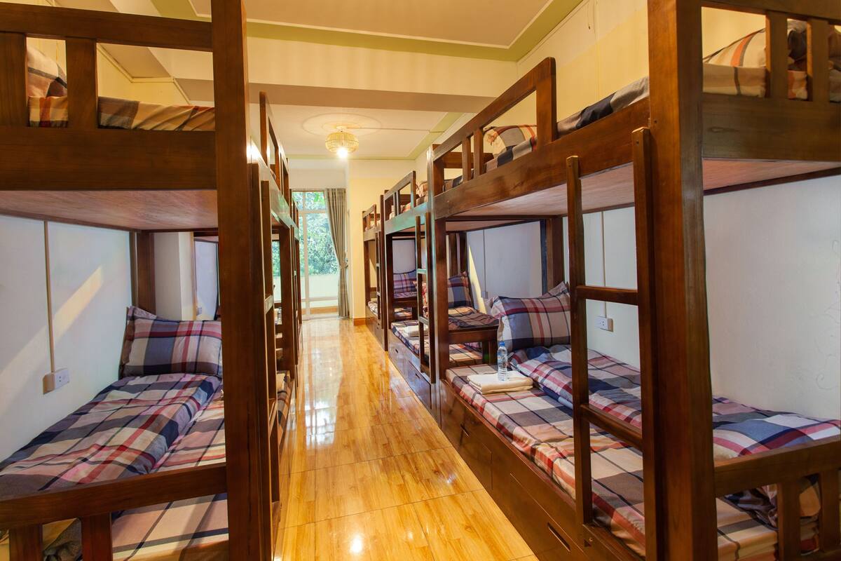 A dormitory room with bunk beds, wooden flooring, and a hallway leading to a door.