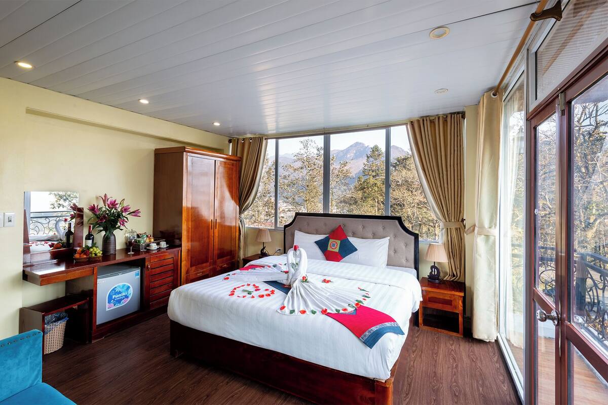 A hotel room with a large bed, wooden furniture, a desk with a computer, and a view of mountains through the window.