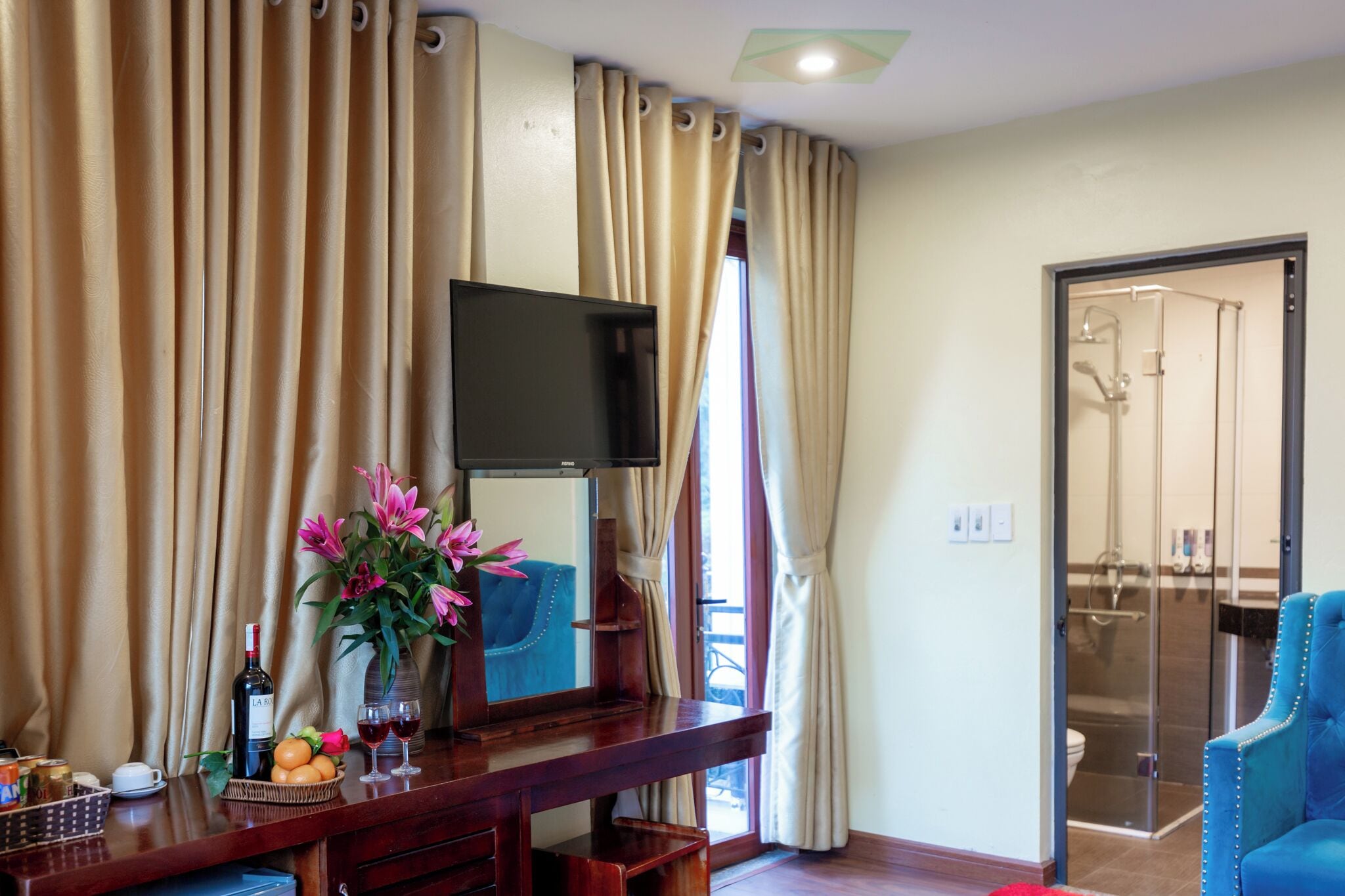 A hotel room with a wooden desk, a flat-screen TV, a vase with flowers, and a blue armchair.