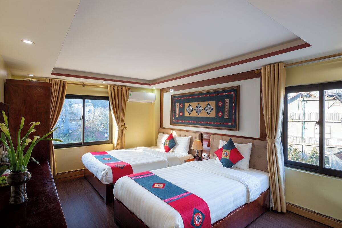 A hotel room with two beds, a large window, a wall-mounted artwork, and a potted plant.