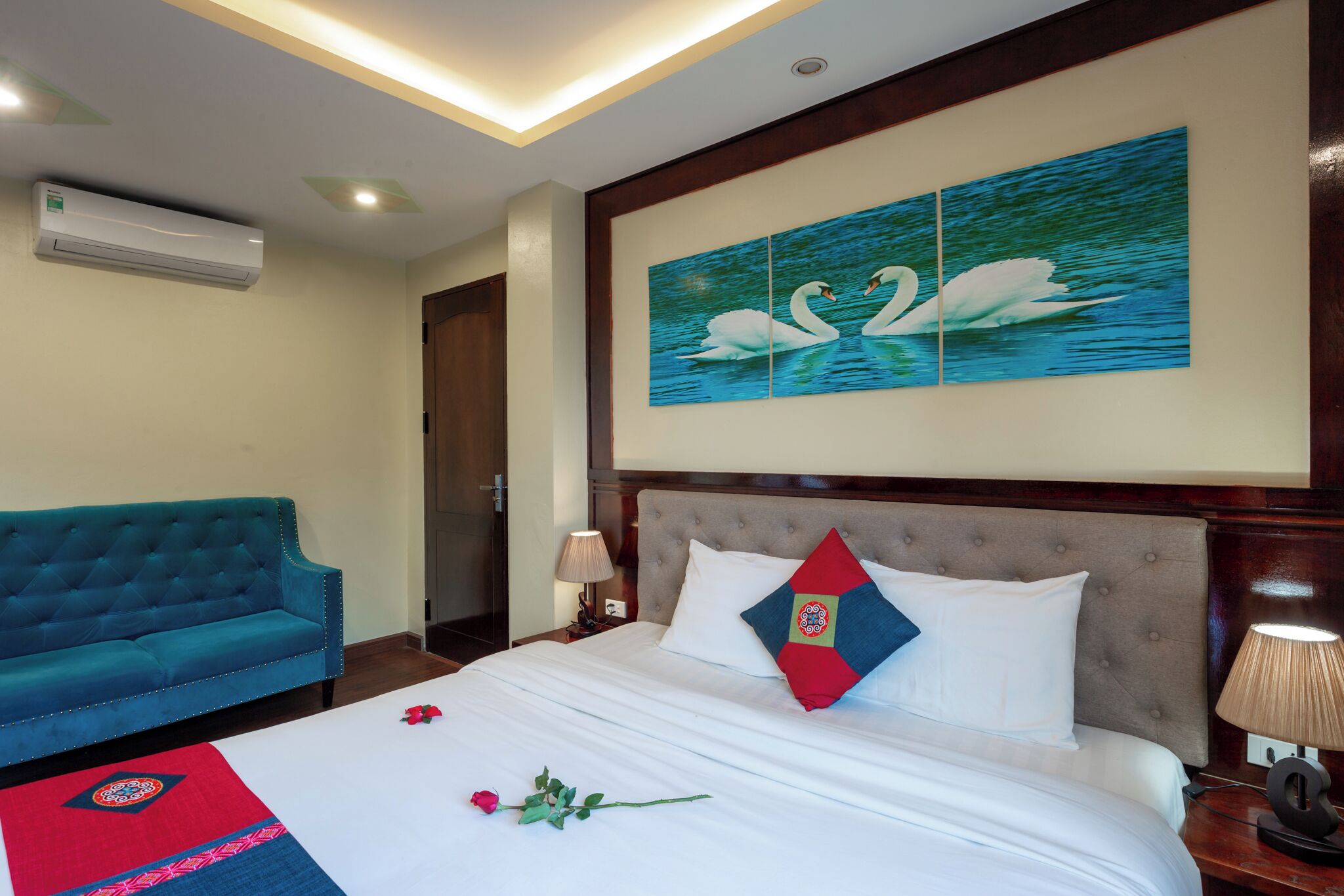 A hotel room with a bed, a blue sofa, a wall with a painting of swans, and an air conditioner.