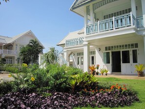 A two-story house with a balcony, surrounded by a well-maintained garden with various plants and flowers. - Moon Margo (Cha-am)