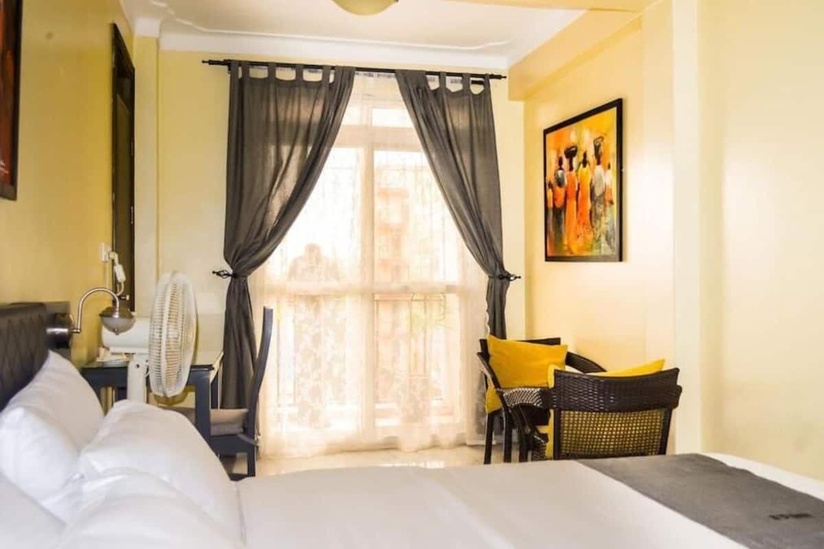 A hotel room with a bed, a desk with a chair, a fan, and a window with curtains.