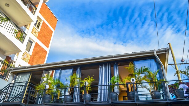 Un hôtel doté d’un balcon orné de plantes en pot, sous un ciel d’un bleu éclatant, et un immeuble à plusieurs étages aux murs rouges et blancs.