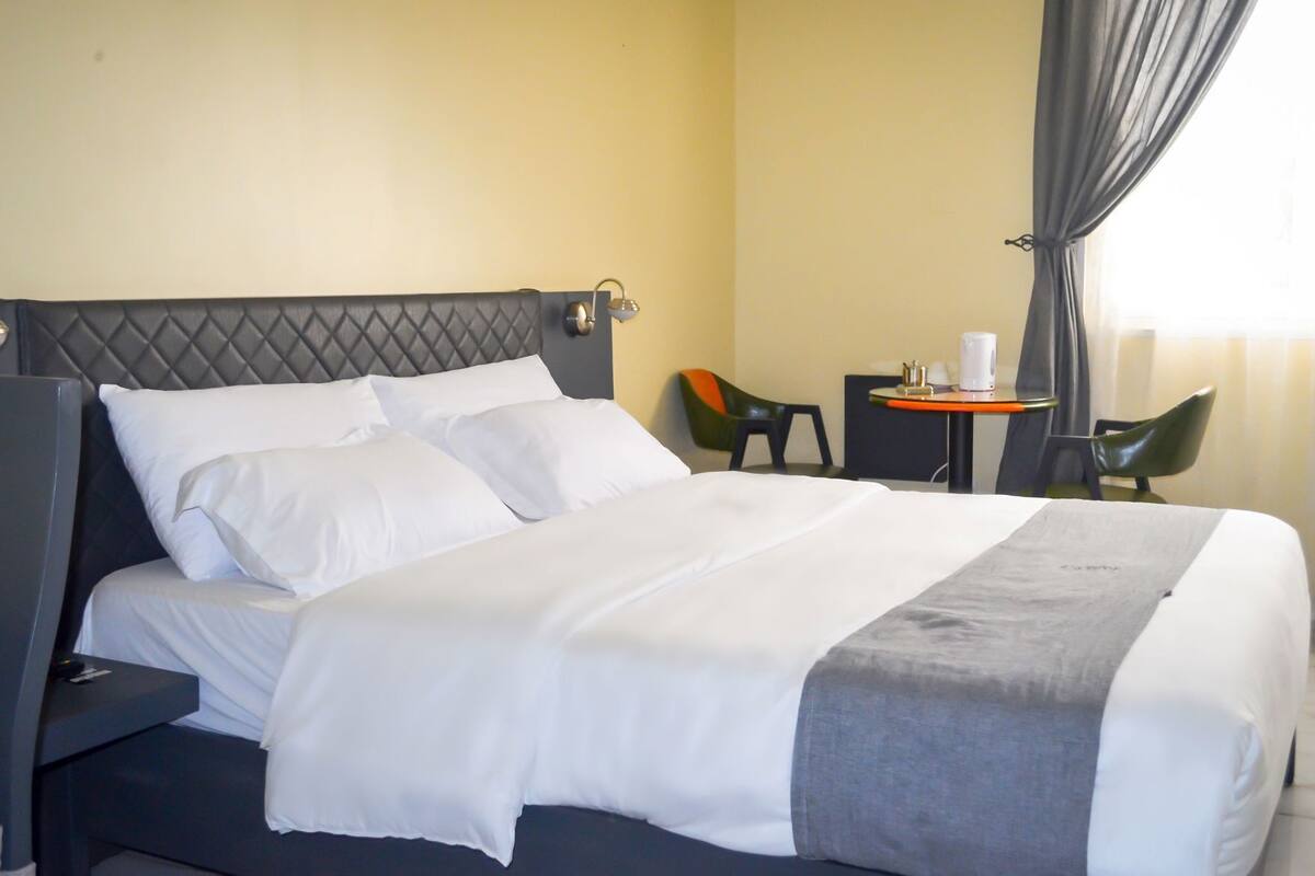 A hotel room with a bed, a small table with a chair, and a window with curtains.