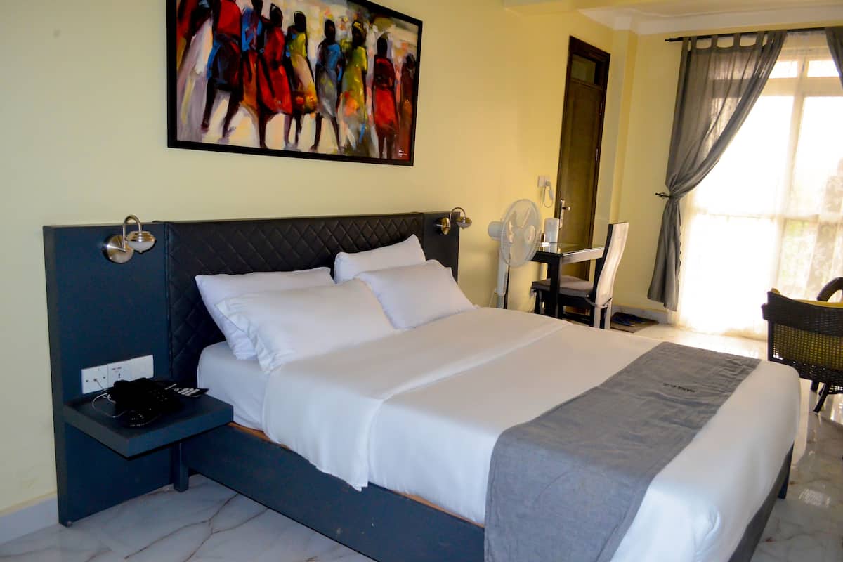 A neatly made bed with white linens, a bedside table with a phone and charger, a wall-mounted artwork, and a window with curtains.