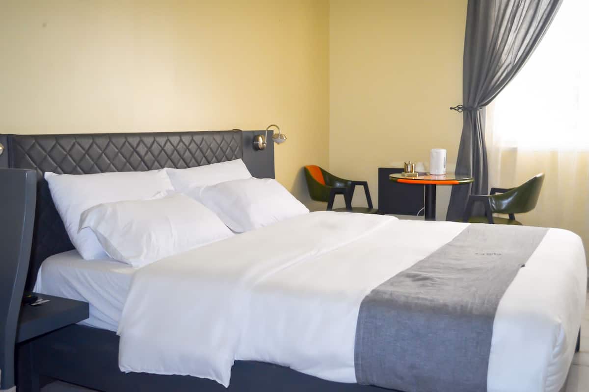 A neatly made bed with white linens and a grey accent, a small table with a white cup and a book, and two chairs in a corner.
