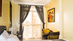 Une chambre d’hôtel avec un lit, un bureau avec une lampe, une chaise avec un coussin jaune, un ventilateur et une fenêtre avec des rideaux légers.