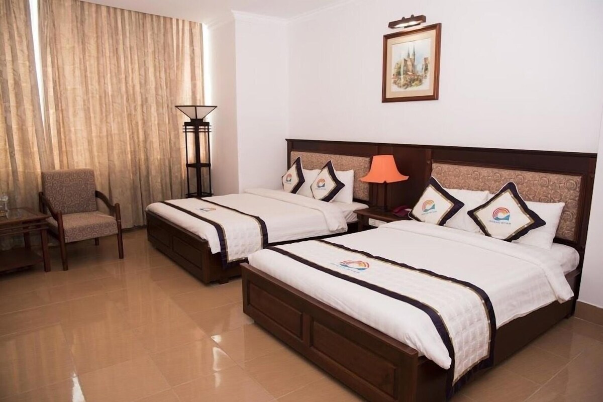 A hotel room with two beds, a chair, a lamp, and a painting on the wall.