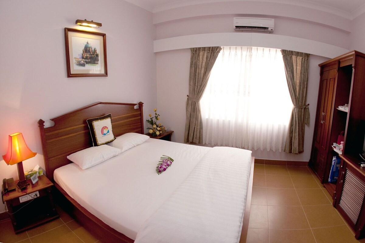 A hotel room with a bed, a nightstand with a lamp, a framed picture on the wall, a window with curtains, and a wardrobe.