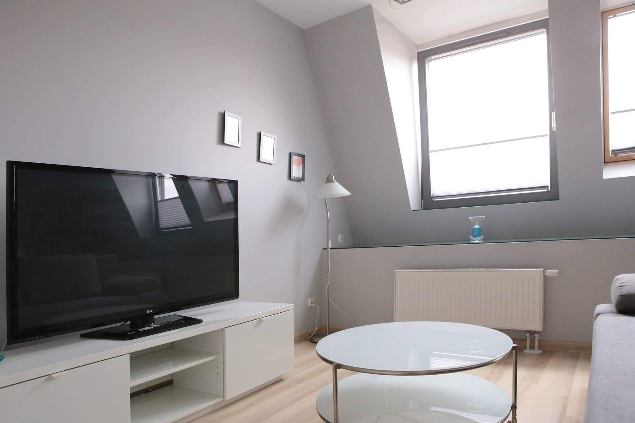 Ein modernes Wohnzimmer mit Fernseher auf einer weißen Unterhaltungseinheit, einem runden Glastisch und einem großen Fenster, das für natürliches Licht sorgt.