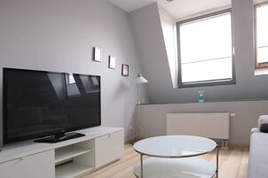 Una sala moderna con un televisor de pantalla plana sobre un mueble blanco, una mesa redonda de vidrio y un ventanal que ofrece luz natural.