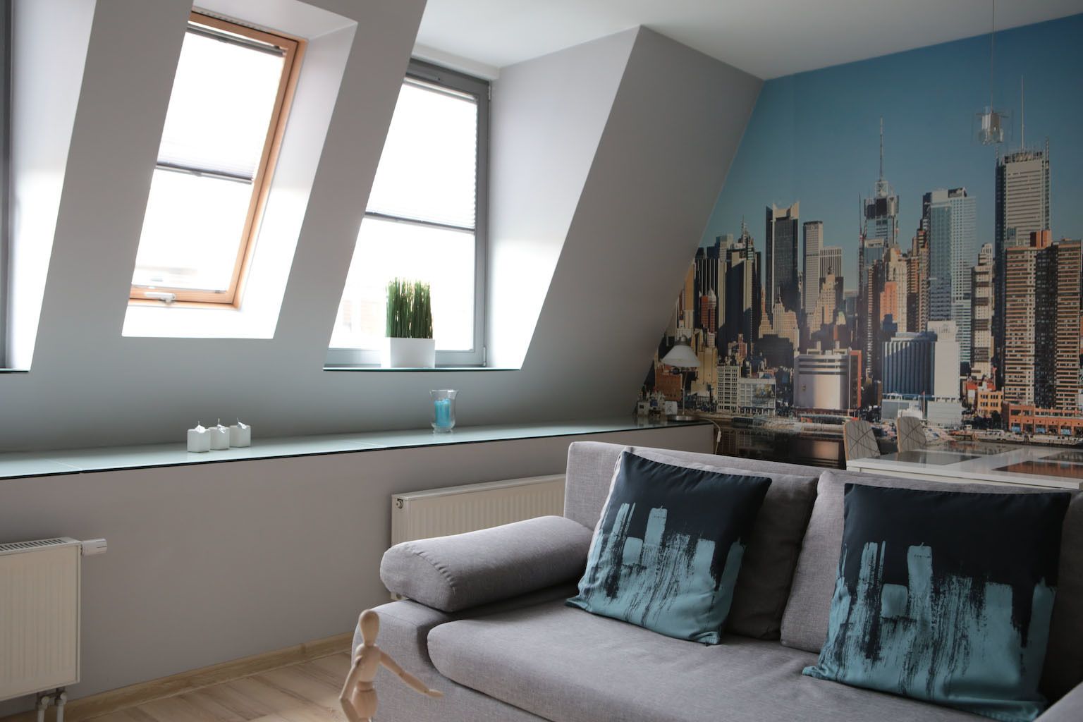 A modern living room with a grey sofa, two windows, a cityscape mural, and a radiator.