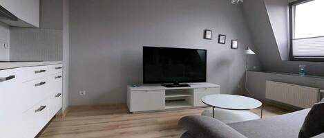 A modern kitchen with white cabinets and a built-in TV. There is a round white table and a grey sofa in the living area.