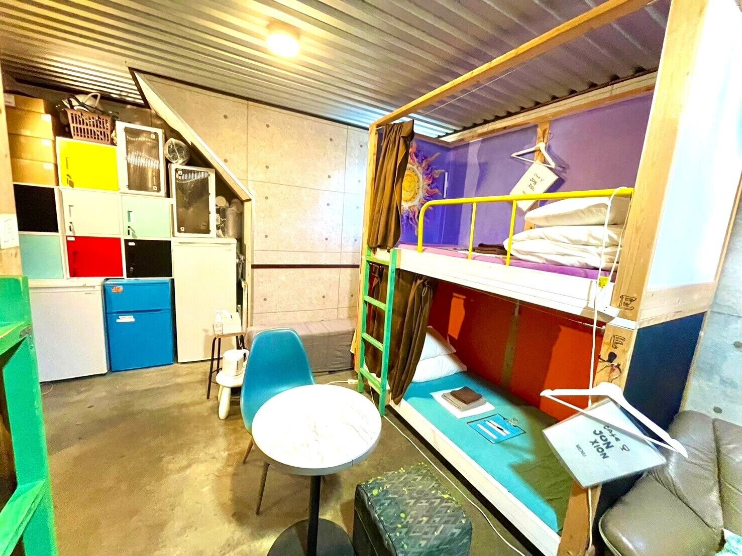 Quarto em dormitório compacto com beliches, uma mesa pequena, uma cadeira e um espaço de cozinha compacta com armários e geladeira.