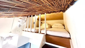 Una habitación pequeña con techo de bambú, una cama con ropa de cama blanca y un ventilador blanco.