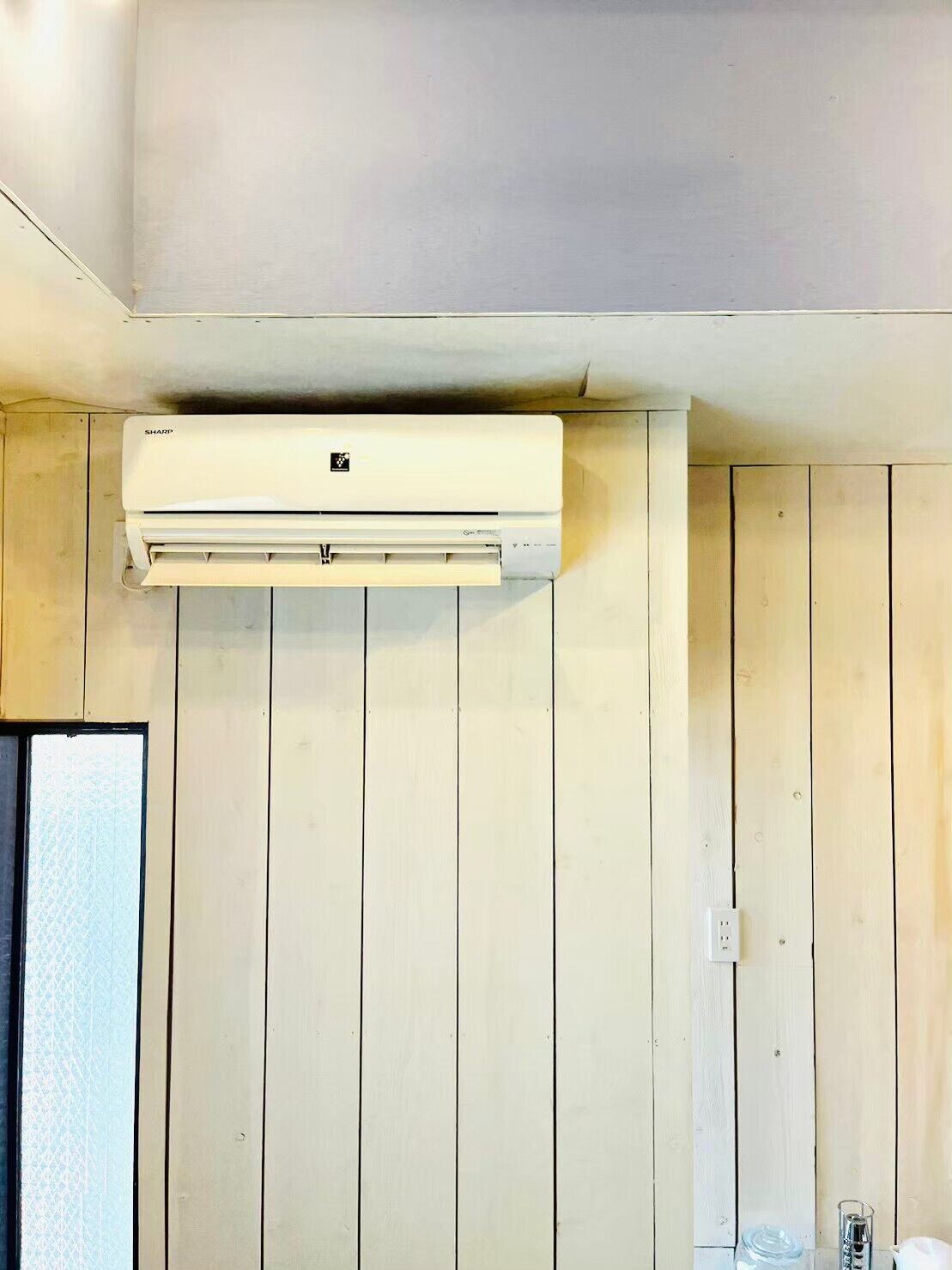Habitació amb parets de fusta, un aparell d'aire acondicionador blanc, una finestra amb vidre mat i una presa elèctrica blanca.