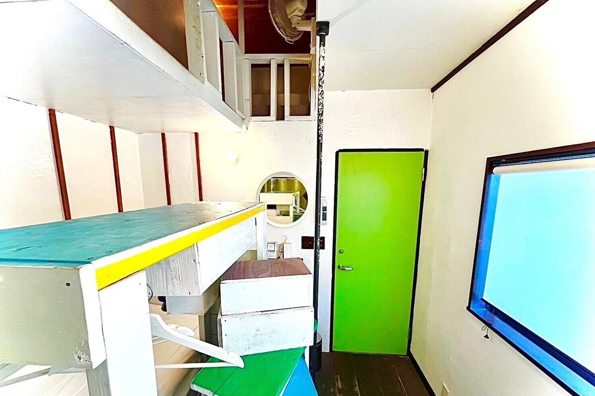 Una habitación estrecha con una cama individual superpuesta, una puerta verde y una ventana.