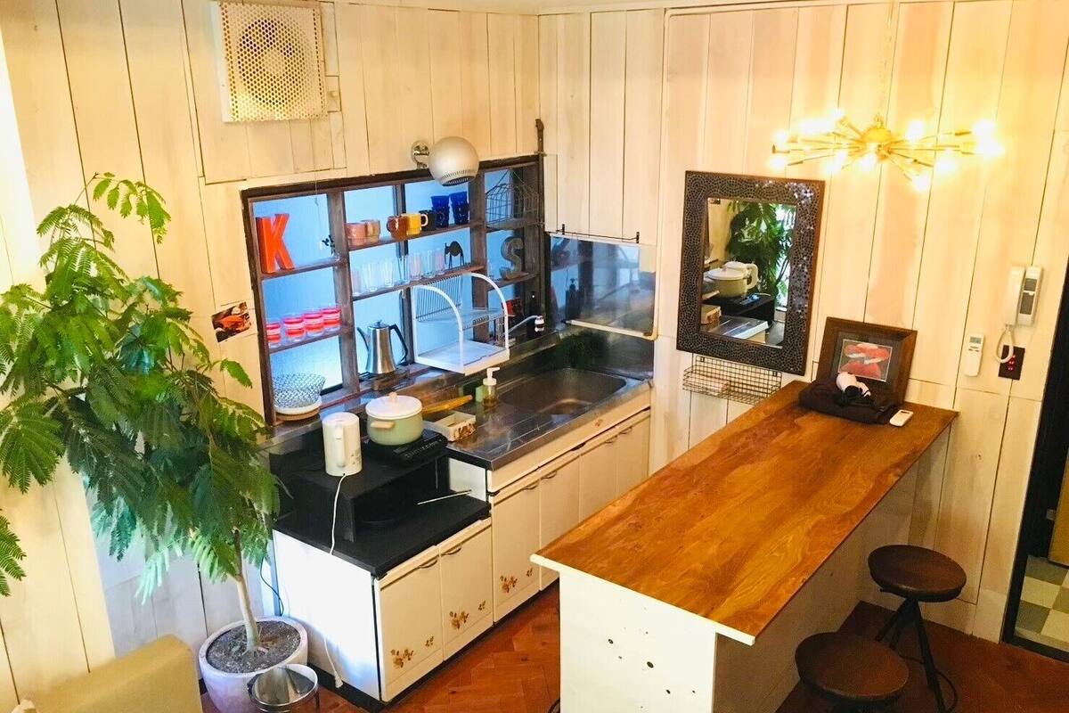 Cocina compacta con mostrador de madera, un lavamanos y un espejo encima. Hay taburetes y una planta en maceta en la habitación.
