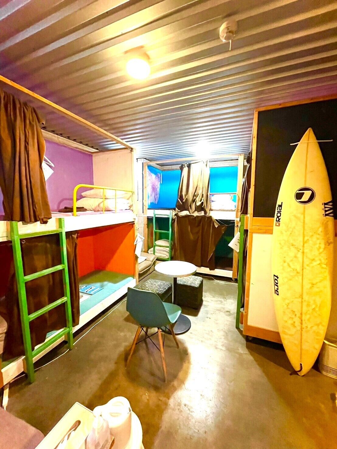 A compact room with bunk beds, a small round table, a chair, and a surfboard.