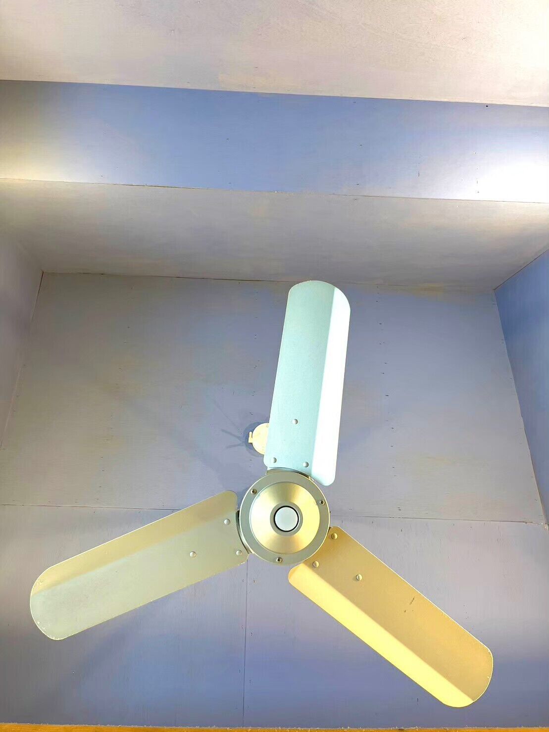 A ceiling fan with three blades in a room with a plain ceiling and light-colored walls.