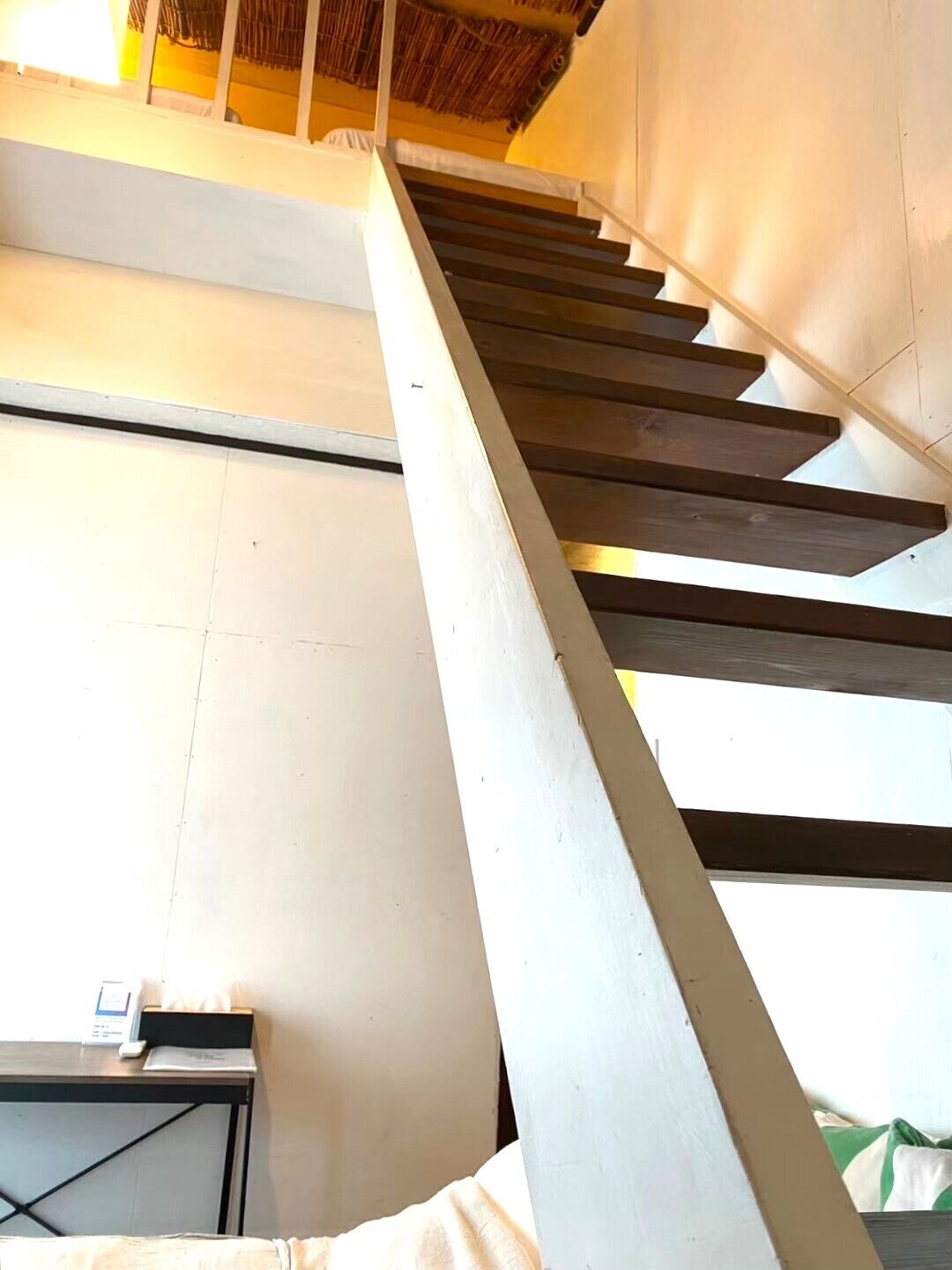A staircase with wooden steps and a white handrail, leading to a room with a bed and a desk.