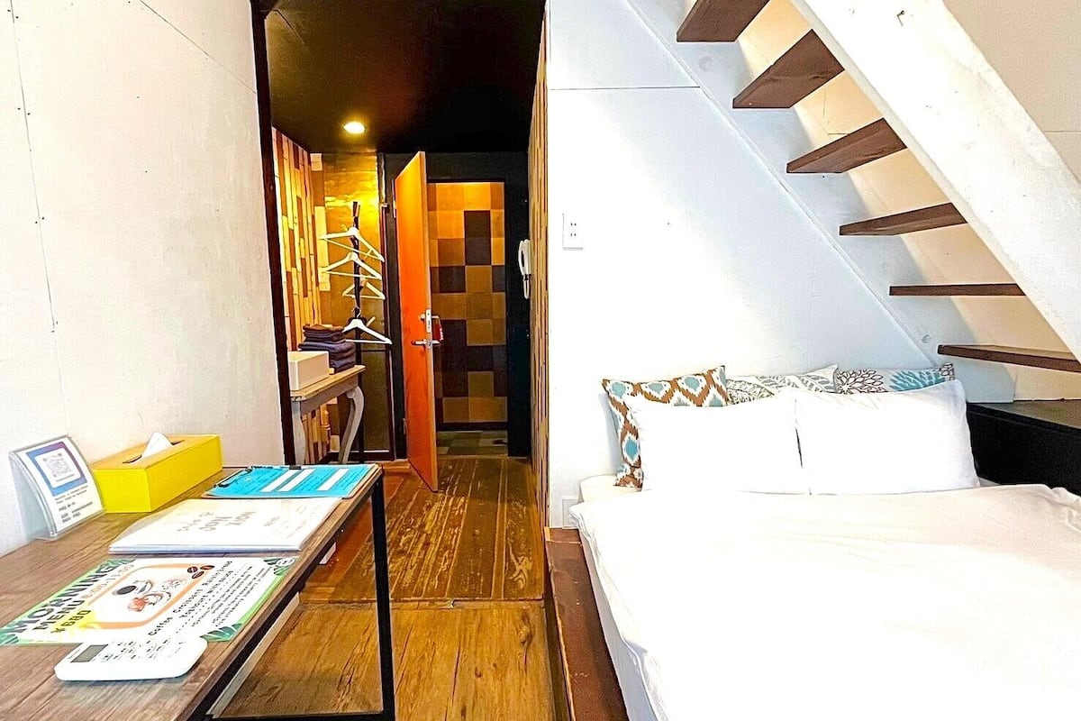 Habitación de hotel con cama, piso de madera, escritorio con silla, perchero y una escalera.
