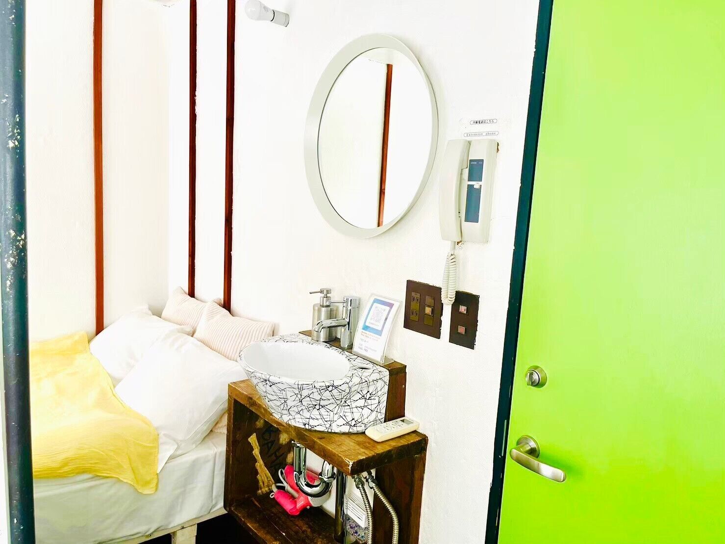 A hotel room with a bed, a bedside table with a mirror, a telephone, and a green door.