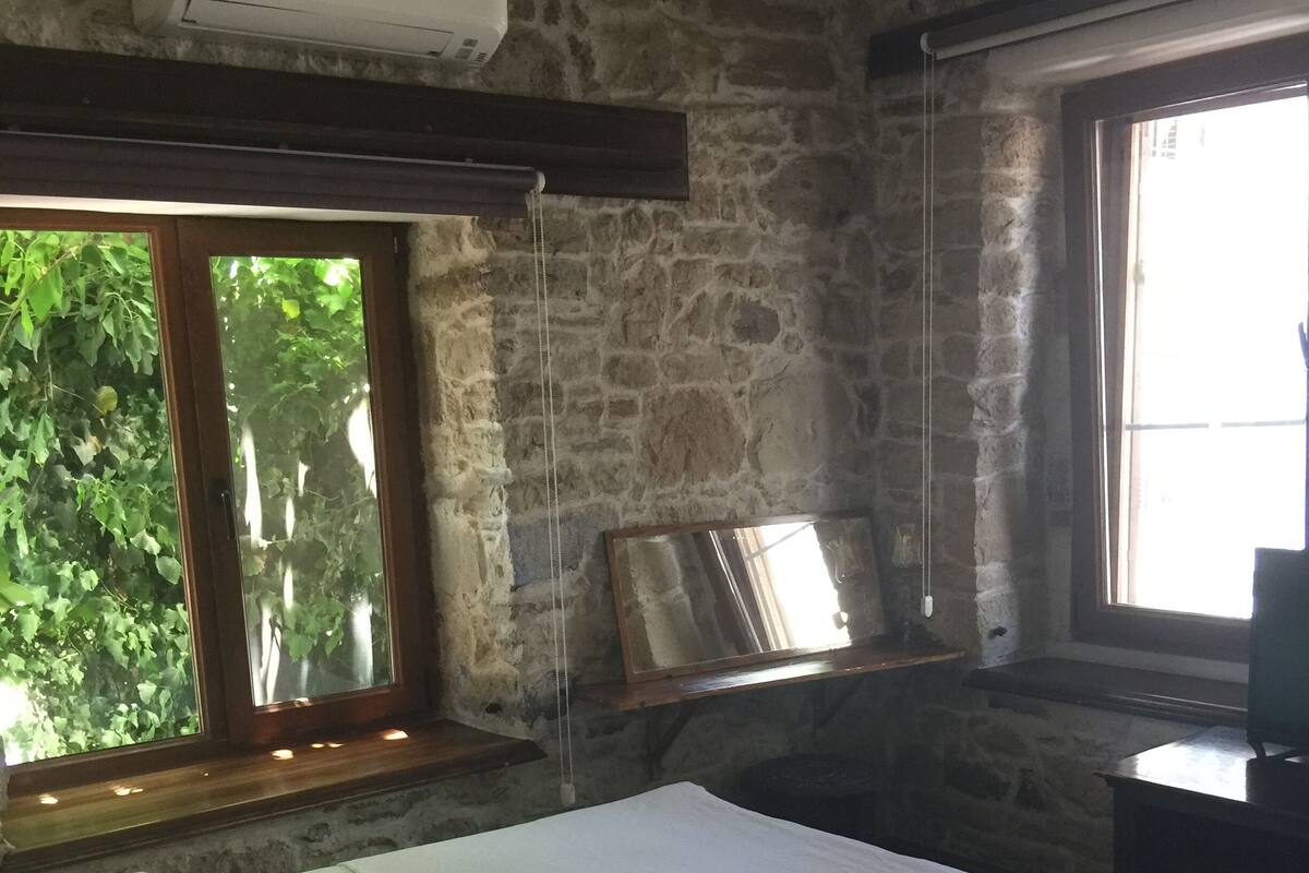 A bedroom with a stone wall, a bed with white bedding, a wooden headboard, a window with a view of greenery, and an air conditioning unit.