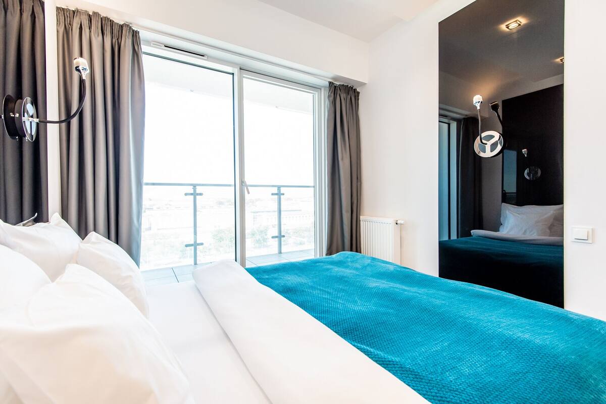 A hotel room with a large bed, a balcony with curtains, a view of the city, and a modern bathroom.