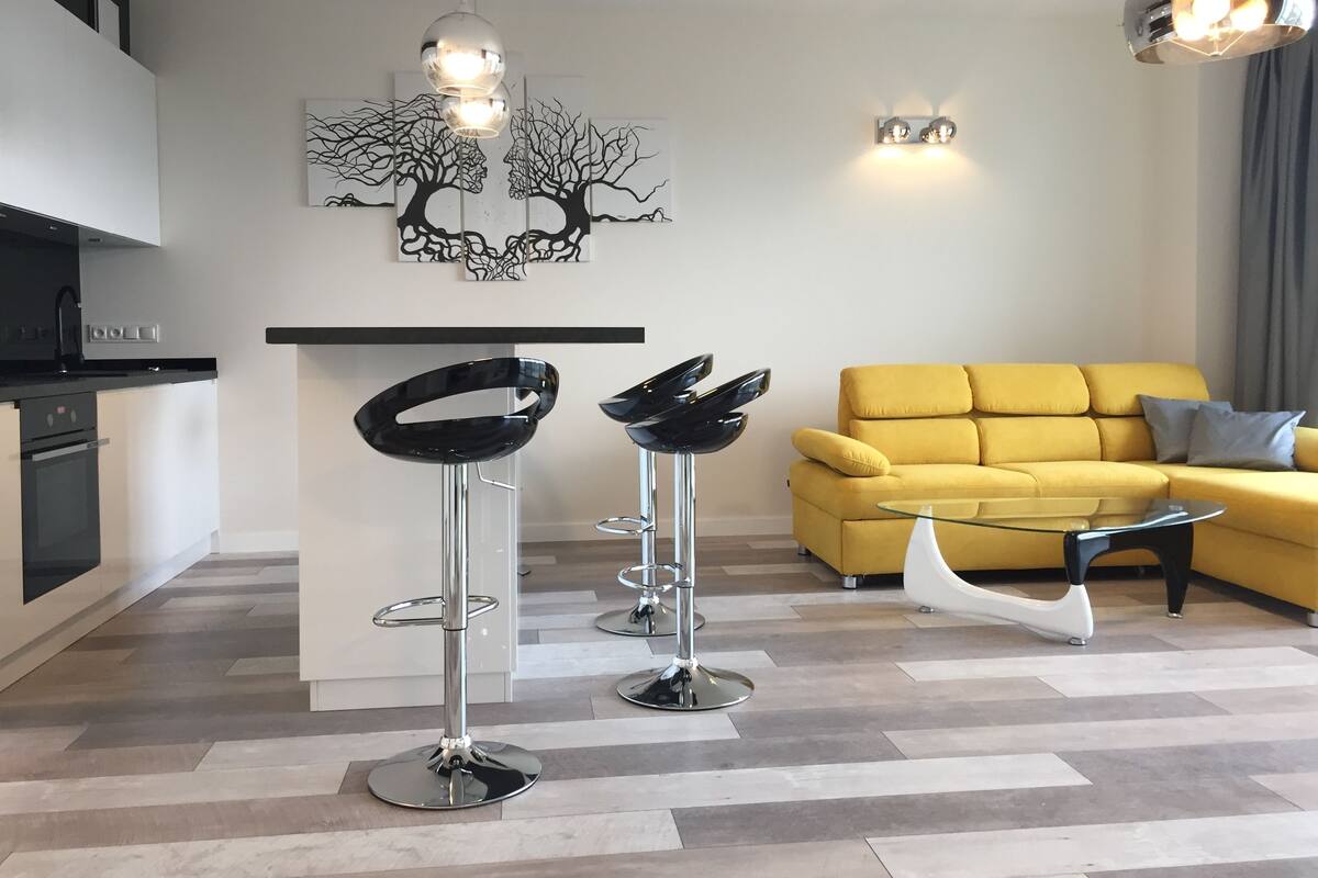 A modern living space with a yellow sofa, two black bar stools, a glass coffee table, and a kitchen area with a black countertop and stainless steel appliances.