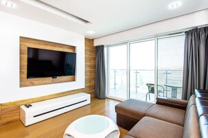 A modern living room with a flat-screen TV mounted on a wooden panel, a white entertainment unit, a glass coffee table, and a brown sofa.