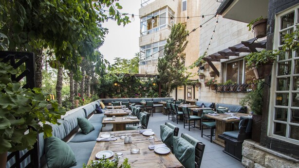 An outdoor dining area with multiple tables and chairs, surrounded by greenery and a building with large windows.
