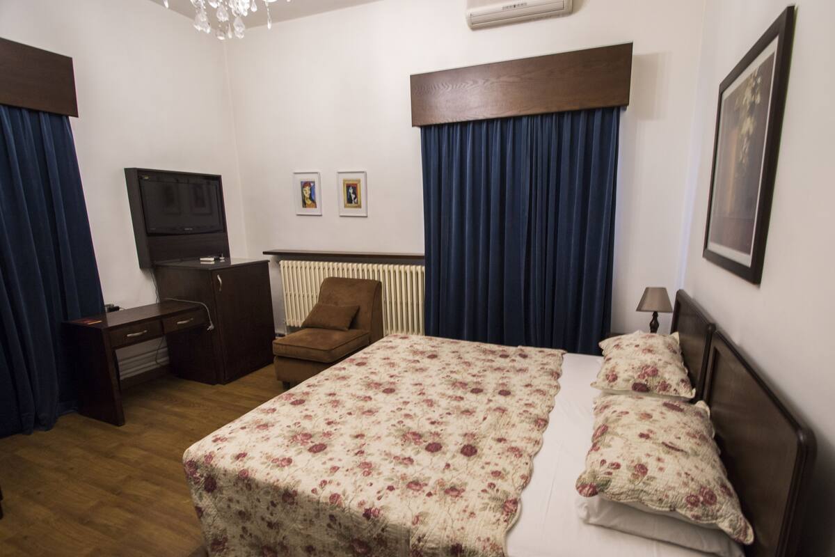 Quarto de hotel com uma cama, escrivaninha, cadeira, televisão e radiador.