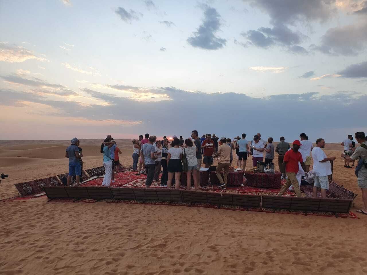 Un grupo de personas reunidas en una playa arenosa, con un bote de madera y una alfombra roja sobre la arena.