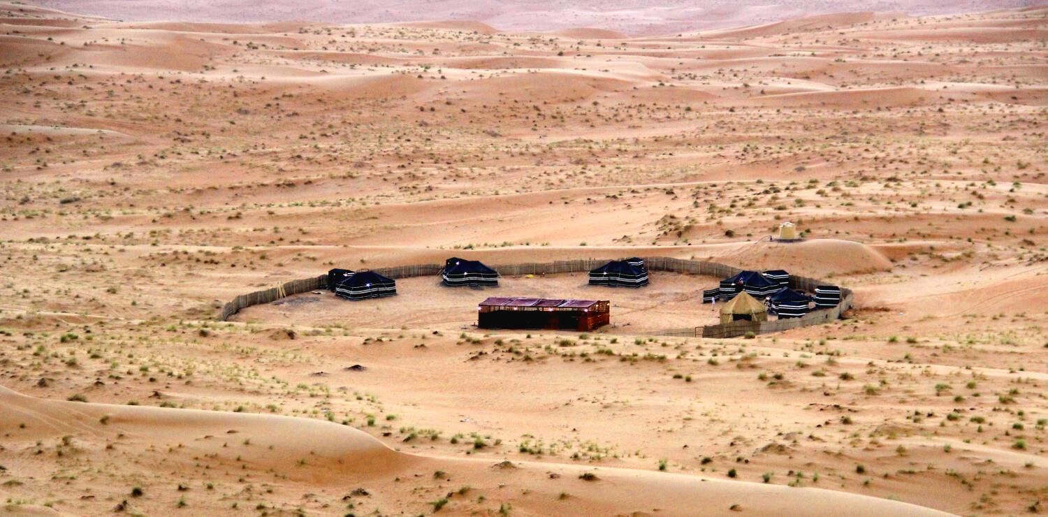 Paisaje desértico con varias tiendas de campaña y un pequeño edificio, probablemente un campamento o alojamiento temporal.