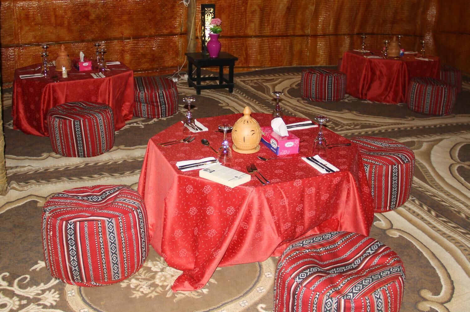 A room with multiple round tables, each covered with a red tablecloth and surrounded by ottomans with patterned cushions.