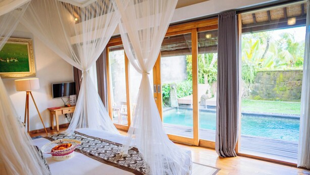 A bedroom with a bed, a television, a bedside table with a lamp, and a view of a swimming pool. - Ume Sri Villas Ubud (Ubud)