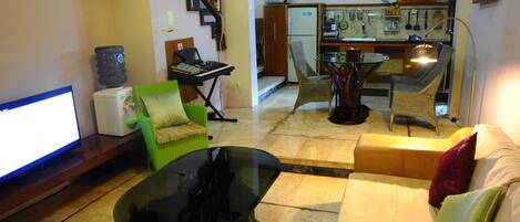 A living room with a sofa, a glass coffee table, a television, a water dispenser, and a kitchen area with a microwave, chairs, and a dining table.