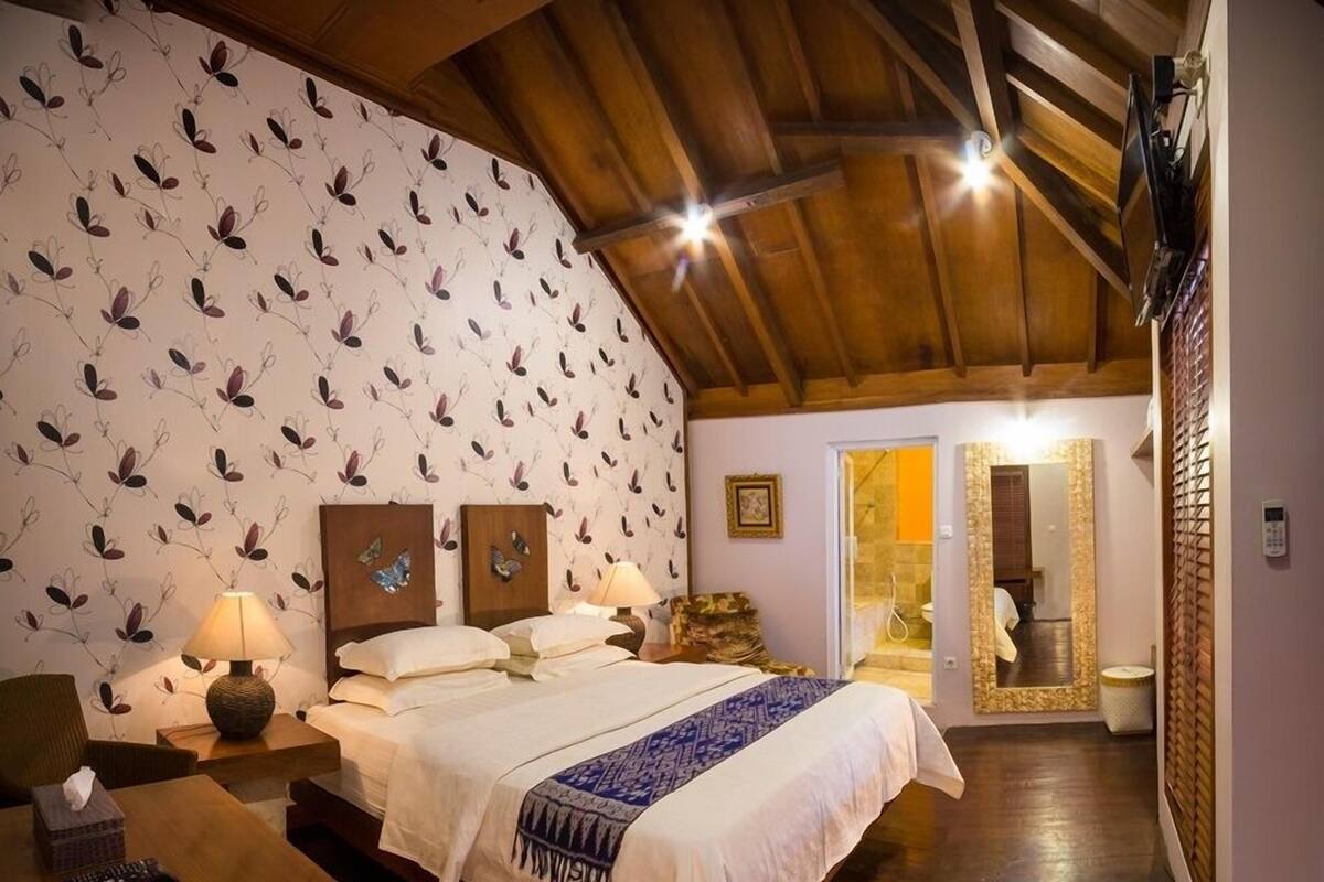 A bedroom with a large bed, wooden headboard, a bedside table with a lamp, and a patterned wallpaper.