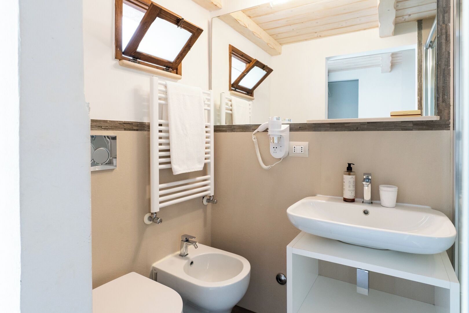 Un baño con inodoro, bidé y dos lavabos. Hay un toallero con una toalla colgada, un espejo sobre el lavabo y una ventana con marco de madera.