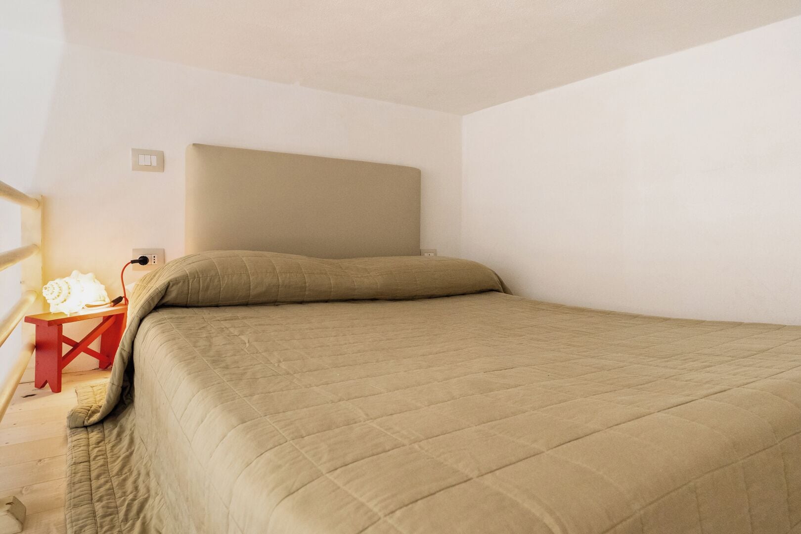 A single bed with a beige quilted bedspread in a room with white walls and a wooden floor.