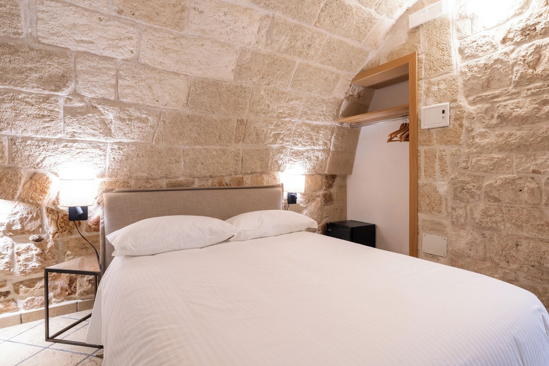 Un dormitorio con pared de piedra, una cama con ropa blanca, cabecero de madera y una mesita de noche.